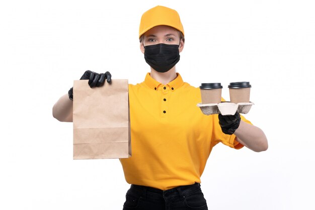 Una joven mensajero de vista frontal en uniforme amarillo guantes negros y máscara negra sosteniendo paquetes de alimentos y tazas de café