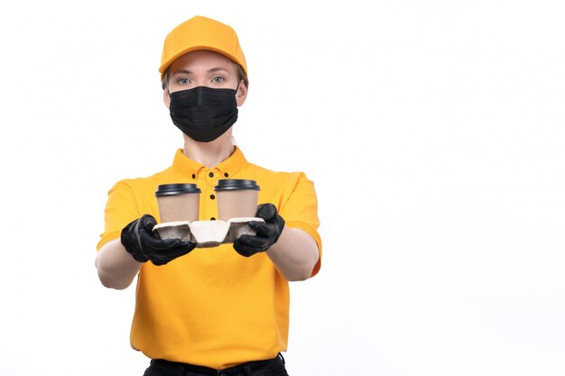 Una joven mensajero de vista frontal en uniforme amarillo guantes negros y máscara negra sosteniendo tazas de café entregando orden