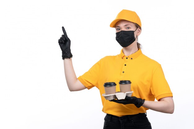 Una joven mensajero de vista frontal en uniforme amarillo guantes negros y máscara negra sosteniendo tazas de café mostrando el signo del dedo