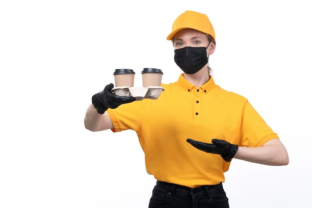 Una joven mensajero de vista frontal en uniforme amarillo guantes negros y máscara negra sosteniendo tazas de café