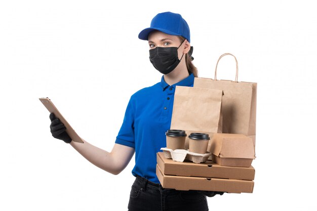 Una joven mensajero de vista frontal en uniforme azul guantes negros y máscara negra con paquetes de entrega de alimentos y bloc de notas