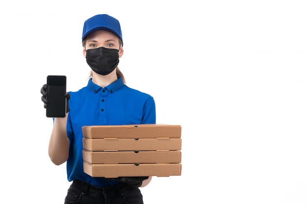 Una joven mensajero de vista frontal en uniforme azul guantes negros y máscara negra con paquetes de entrega de alimentos y teléfono inteligente