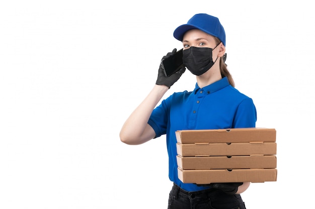 Una joven mensajero de vista frontal en uniforme azul guantes negros y máscara negra con paquetes de entrega de alimentos y teléfono inteligente