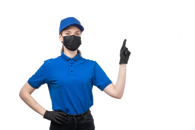 Una joven mensajero de vista frontal en uniforme azul guantes negros y máscara negra posando y mostrando el signo señalador