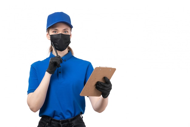 Una joven mensajero de vista frontal en uniforme azul guantes negros y máscara negra sosteniendo el bloc de notas