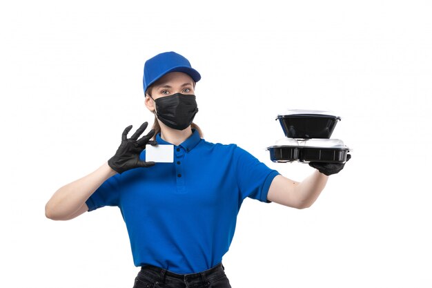 Una joven mensajero de vista frontal en uniforme azul guantes negros y máscara negra sosteniendo cuencos de entrega de alimentos y tarjeta blanca