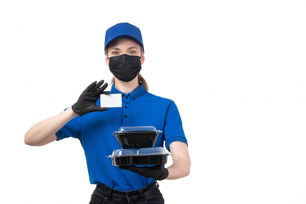 Una joven mensajero de vista frontal en uniforme azul guantes negros y máscara negra sosteniendo cuencos de entrega de alimentos y tarjeta blanca