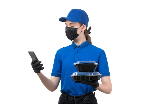 Una joven mensajero de vista frontal en uniforme azul guantes negros y máscara negra sosteniendo cuencos de entrega de alimentos y teléfono