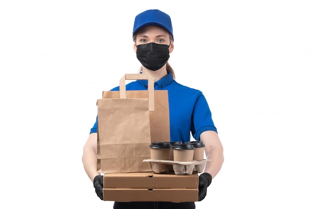 Una joven mensajero de vista frontal en uniforme azul guantes negros y máscara negra sosteniendo paquetes de entrega de alimentos