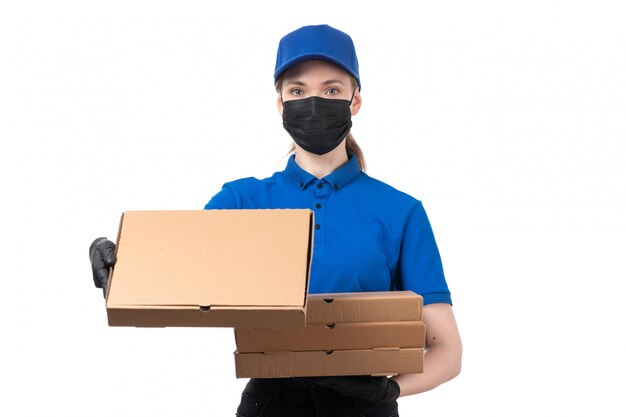 Una joven mensajero de vista frontal en uniforme azul guantes negros y máscara negra sosteniendo paquetes de entrega de alimentos