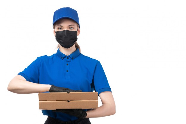 Una joven mensajero de vista frontal en uniforme azul guantes negros y máscara negra sosteniendo paquetes de entrega de alimentos