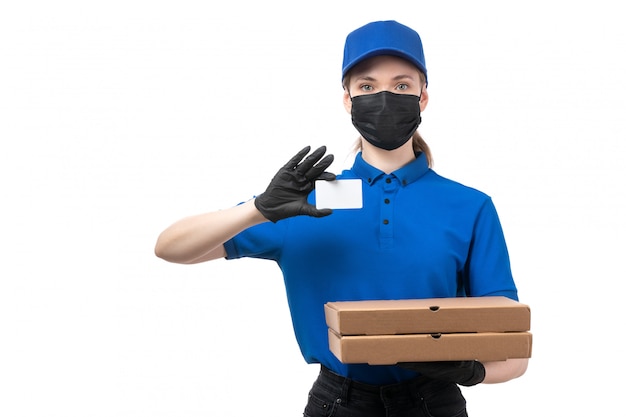 Una joven mensajero de vista frontal en uniforme azul guantes negros y máscara negra sosteniendo paquetes de entrega de alimentos