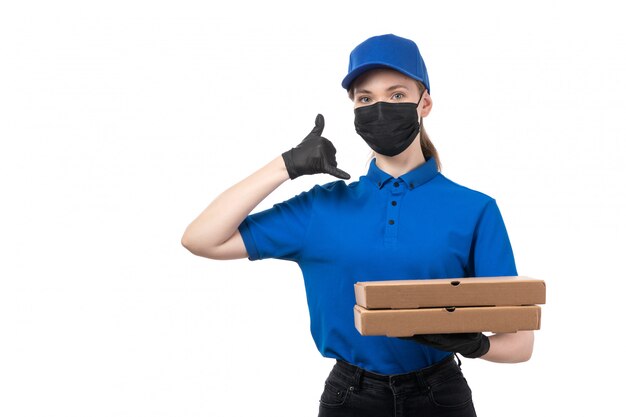 Una joven mensajero de vista frontal en uniforme azul guantes negros y máscara negra sosteniendo paquetes de entrega de alimentos