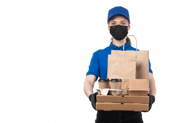 Una joven mensajero de vista frontal en uniforme azul guantes negros y máscara negra sosteniendo paquetes de entrega de alimentos