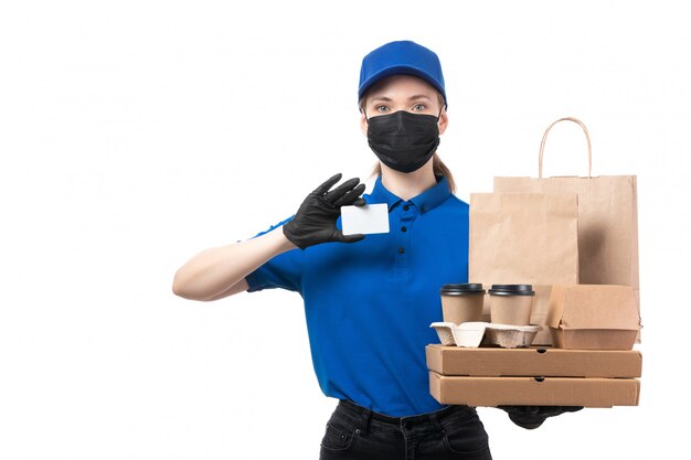 Una joven mensajero de vista frontal en uniforme azul guantes negros y máscara negra sosteniendo paquetes de entrega de alimentos