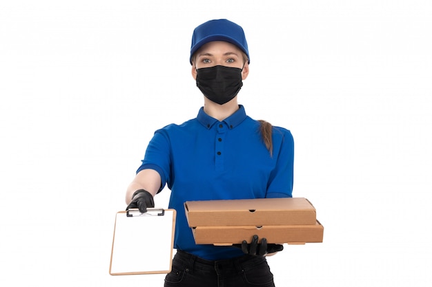 Una joven mensajero de vista frontal en uniforme azul máscara negra y guantes con paquetes de entrega de alimentos y bloc de notas