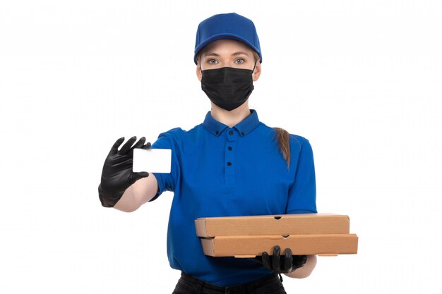 Una joven mensajero de vista frontal en uniforme azul máscara negra y guantes con paquetes de entrega de alimentos y tarjeta blanca