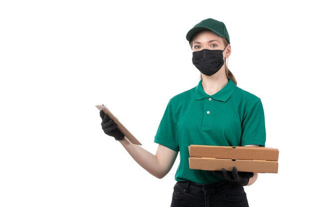 Una joven mensajero de vista frontal en uniforme verde guantes negros y máscara negra con cajas de entrega de alimentos