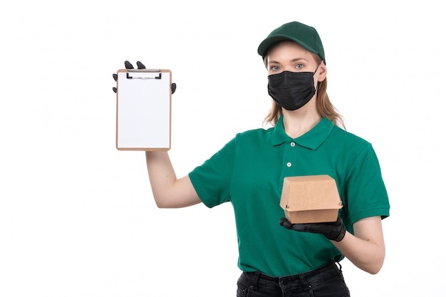 Una joven mensajero de vista frontal en uniforme verde guantes negros y máscara negra con paquete de entrega de alimentos y bloc de notas