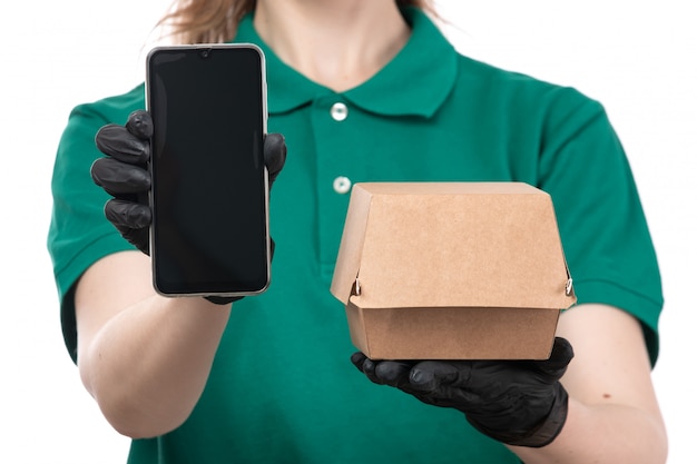 Una joven mensajero de vista frontal en uniforme verde guantes negros y máscara negra con paquete de entrega de alimentos y teléfono