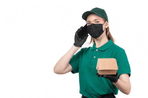Una joven mensajero de vista frontal en uniforme verde guantes negros y máscara negra con paquete de entrega de alimentos y teléfono