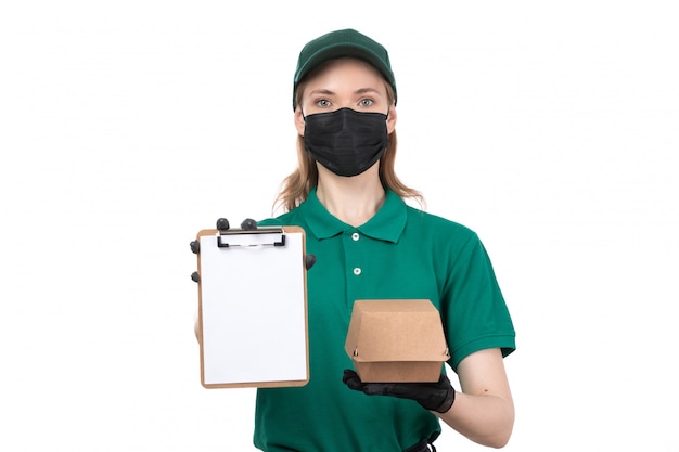 Una joven mensajero de vista frontal en uniforme verde guantes negros y máscara negra con paquete de entrega de alimentos