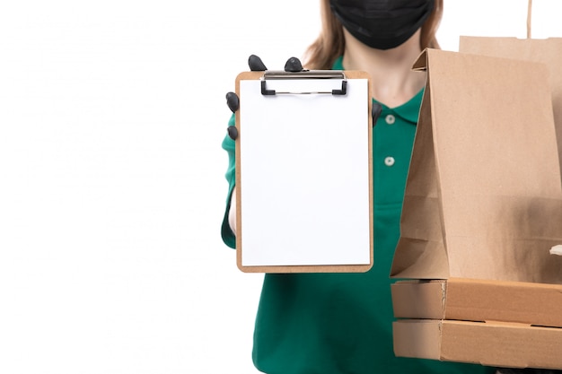 Una joven mensajero de vista frontal en uniforme verde guantes negros y máscara negra sosteniendo paquetes de entrega de alimentos y entrega de bloc de notas