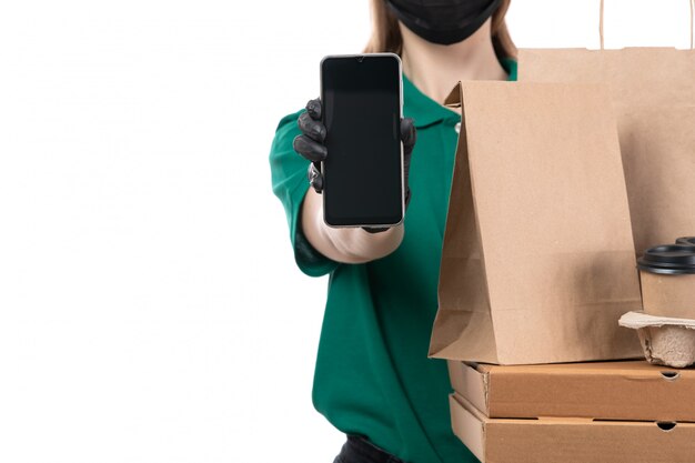 Una joven mensajero de vista frontal en uniforme verde guantes negros y máscara negra sosteniendo paquetes de entrega de alimentos y entrega de teléfono