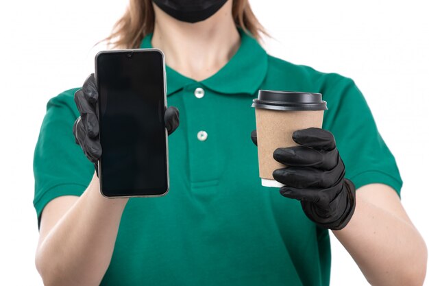 Una joven mensajero de vista frontal en uniforme verde guantes negros y máscara negra sosteniendo la taza de café y el teléfono