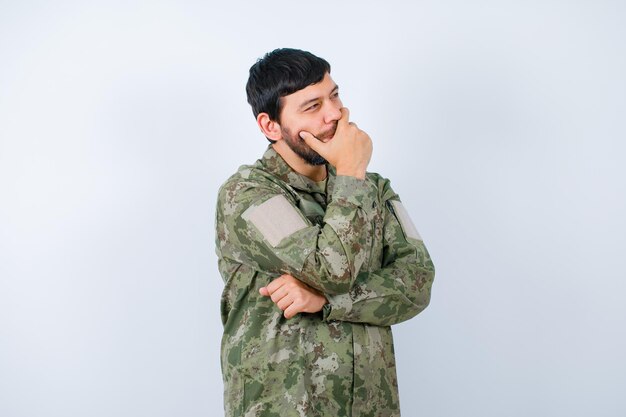 El joven militar está pensando tomándose la mano en la boca y mirando hacia otro lado en el fondo blanco