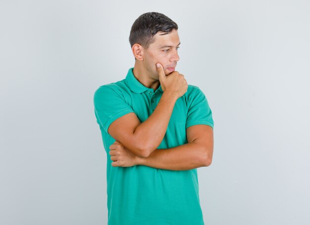 Joven mirando al lado con la mano en la barbilla en camiseta verde y mirando pensativo. vista frontal.