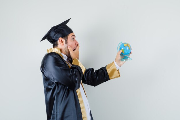 Joven mirando el globo de la escuela en uniforme de posgrado y mirando sorprendido.