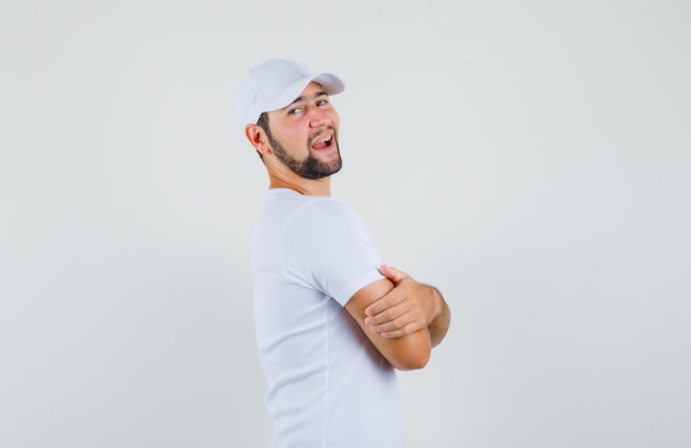 Joven mirando a un lado mientras está de pie con los brazos cruzados en camiseta, gorra y mirando feliz.