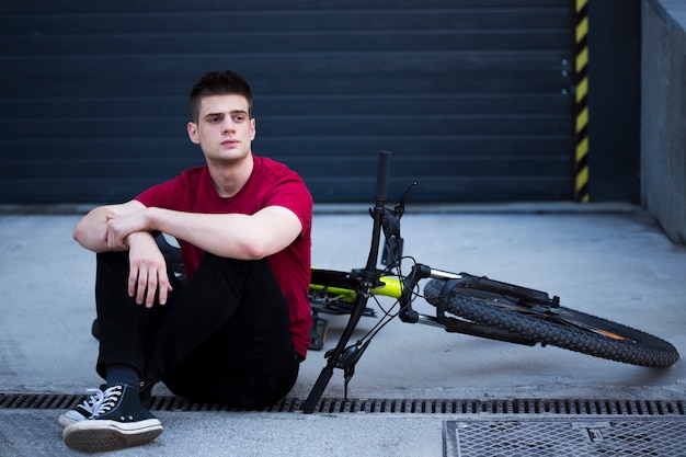 Foto gratuita joven mirando a otro lado y sentado junto a la bicicleta