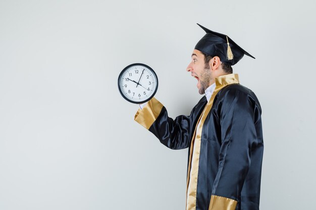 Joven mirando el reloj de pared en uniforme de posgrado y mirando feliz. .