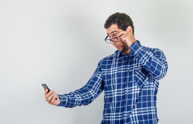 Joven mirando smartphone sobre gafas en camisa a cuadros y mirando consternado