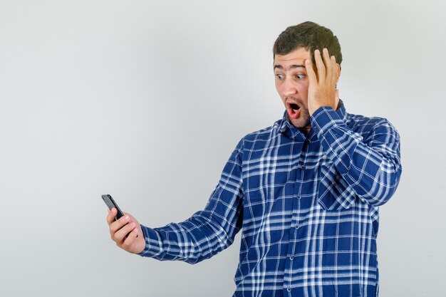 Joven mirando el teléfono inteligente con la mano en la cabeza en camisa a cuadros y mirando sorprendido