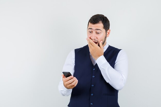 Joven mirando el teléfono móvil en camisa, chaleco y mirando asombrado