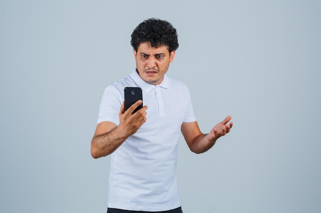 Joven mirando el teléfono móvil en camiseta blanca y mirando confundido, vista frontal.