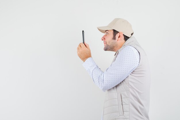 Joven mirando el teléfono móvil con chaqueta y gorra beige y mirando feliz, vista frontal.