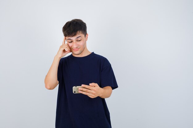 Joven mirando el teléfono móvil, manteniendo la mano en la cabeza con una camiseta negra y mirando pensativo, vista frontal.