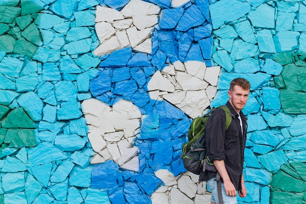 Joven con mochila de viaje de pie cerca de la pared de piedra pintada