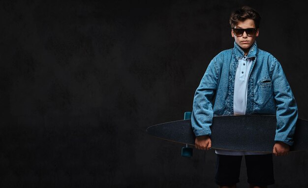 Un joven de moda con gafas de sol vestido con una camiseta y una chaqueta vaquera sostiene un longboard. Aislado en el fondo oscuro.