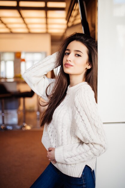 Joven modelo hermosa con labios rojos y cabello largo y castaño en suéter blanco y jeans azules, se queda en la habitación de su casa