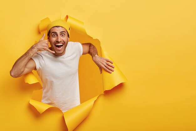 Joven modelo masculino atractivo positivo hace gesto de llamada, viste sombrero amarillo y camiseta blanca informal