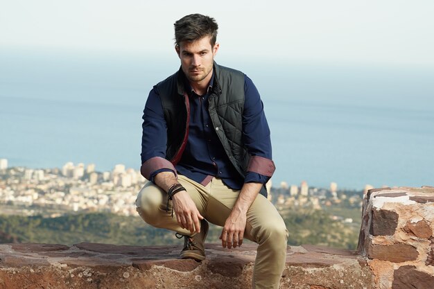 Joven modelo masculino hispano vistiendo una camisa azul y una chaqueta negra posando cerca de un muro de piedra