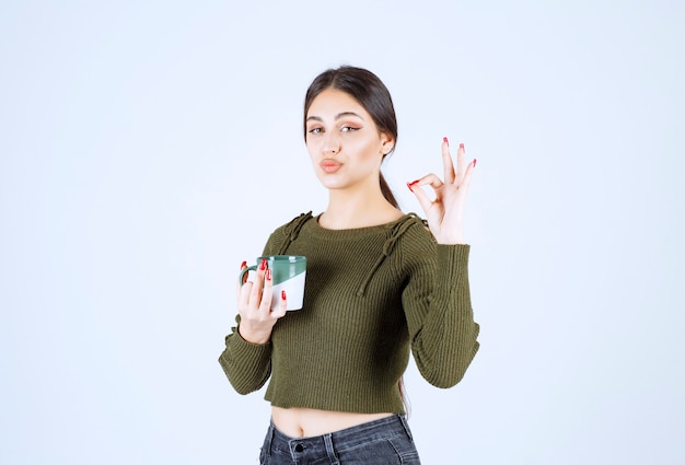 Un joven modelo de mujer sonriente sosteniendo una taza y mostrando un gesto bien.