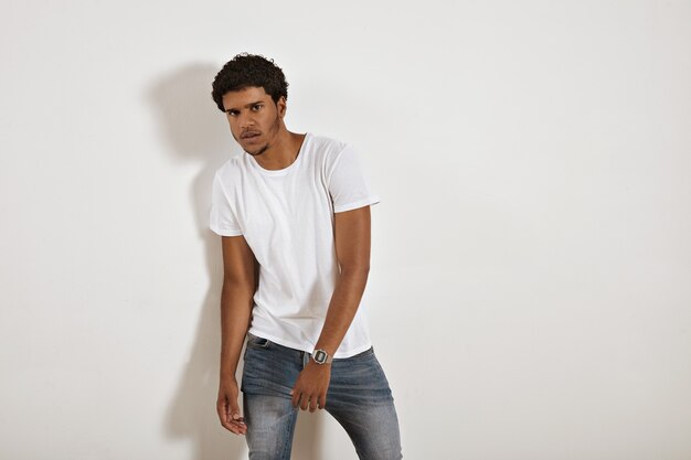 Joven modelo negro de aspecto serio en jeans y camiseta blanca en blanco en la pared blanca