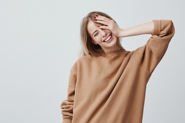 La joven modelo rubia atractiva y positiva usa un suéter informal suelto, y está feliz de recibir buenas noticias. Mujer alegre se regocija los fines de semana, relajándose en el interior