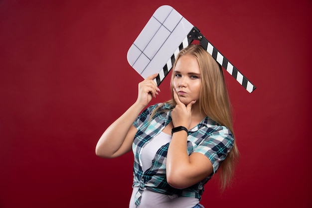 Foto gratuita joven modelo rubia sosteniendo un tablero de azote de filmación de películas en blanco y parece confundido.